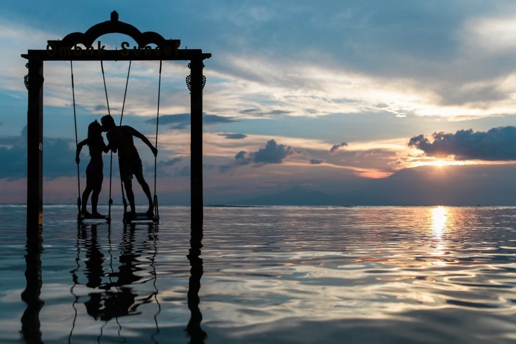 Silhouette of two lovers standing on a giant swing in the sea at sunset